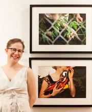 Dr. 伊凡娜乔治 smiling with brown hair pulled up wearing black rim glasses and a sleeveless v-neck off white dress standing beside 2 framed photographs on display