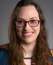 Rachael Linker smiling with brown rimmed glasses and long wavy brown hair and wearing a multicolored blouse