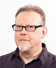 headshot photo of Tony Schultz with receding light brown hair and graying beard and mustache wearing thin black rim glasses and a brown button down shirt